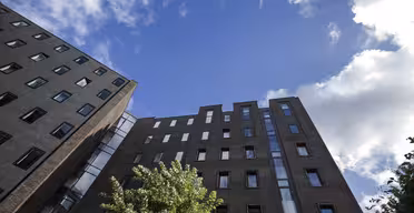 Uppåtgående vy av två moderna tegelbyggnader med flera fönster mot en blå himmel, utspridda med moln. Ett lummigt träd är synligt längst ner, vilket ger en touch av grönska till den urbana scenen.