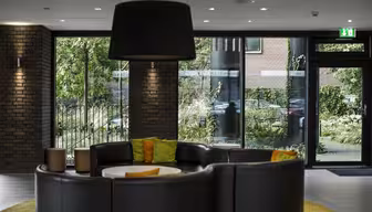 Modern lobby med en rund svart skinnsoffa och färgglada kuddar. Stora fönster ger utsikt över frodig grönska utanför. En stor svart taklampa hänger över huvudet, och tegelväggar ger en stilfull kontrast till den eleganta inredningen.