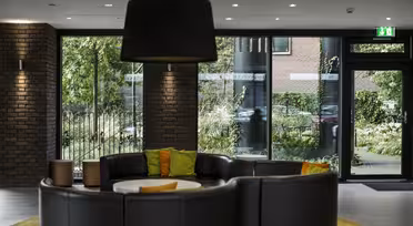 Modern lobby med en rund svart skinnsoffa och färgglada kuddar. Stora fönster ger utsikt över frodig grönska utanför. En stor svart taklampa hänger över huvudet, och tegelväggar ger en stilfull kontrast till den eleganta inredningen.