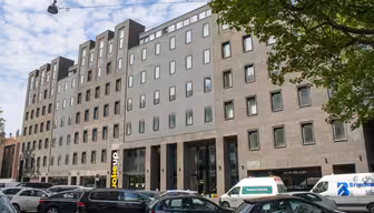 En modern sexvåningsbyggnad med en blandning av ljusgrå och tegelfasad har många fönster. Det står bilar parkerade längs gatan framför, och träd ramar delvis in bilden. Himlen är delvis molnig.