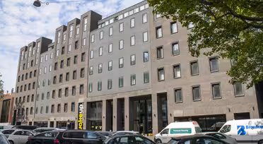 En modern sexvåningsbyggnad med en blandning av ljusgrå och tegelfasad har många fönster. Det står bilar parkerade längs gatan framför, och träd ramar delvis in bilden. Himlen är delvis molnig.