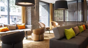 Modern hotellobby med stora fönster, grå soffor som accentueras av gröna och orangea kuddar, läderstolar, runda bord och stora mörka taklampor. Lokalen är fylld med naturligt ljus och har en mysig och inbjudande atmosfär.