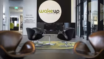 Modern hotellobby med fyra bruna snurrstolar runt runda bord på en grönmönstrad matta. En stor svart och vit vägg har logotypen "Wakeup Copenhagen". Glasdörrar och en hall syns i bakgrunden.
