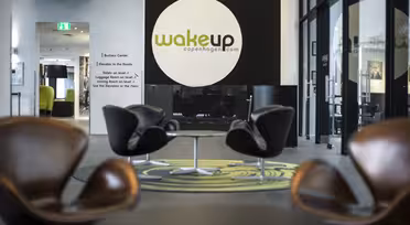 Modern hotellobby med fyra bruna snurrstolar runt runda bord på en grönmönstrad matta. En stor svart och vit vägg har logotypen "Wakeup Copenhagen". Glasdörrar och en hall syns i bakgrunden.