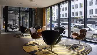 Ett modernt kafé med stora fönster har svarta och bruna snurrstolar på en mönstrad matta. Utanför står bilar och en cykel på gatan med byggnader i bakgrunden. Atmosfären är lätt och luftig.