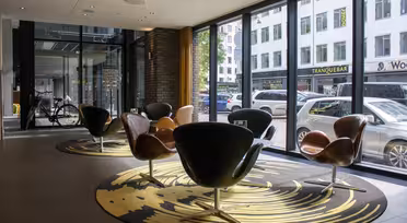 Ett modernt kafé med stora fönster har svarta och bruna snurrstolar på en mönstrad matta. Utanför står bilar och en cykel på gatan med byggnader i bakgrunden. Atmosfären är lätt och luftig.