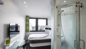 Ett modernt hotellrum har en säng vid ett fönster, ett duschvägg i glas, en platt-TV på väggen och ett genomskinligt skrivbord i glas med en stol. Rummet är ljust med naturligt ljus från stora fönster.