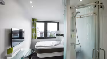 Ett modernt hotellrum har en säng vid ett fönster, ett duschvägg i glas, en platt-TV på väggen och ett genomskinligt skrivbord i glas med en stol. Rummet är ljust med naturligt ljus från stora fönster.