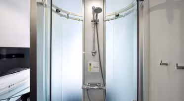 Modern glasdusch med svängda skjutdörrar och silverbeslag. Inuti är ett duschmunstycke monterat på en central panel tillsammans med en tvålautomat. Badrummet har ljusa väggar och en krok till höger.