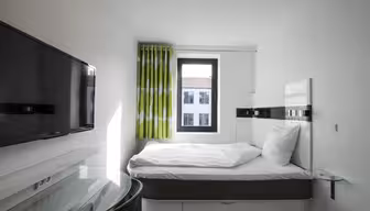 Ett kompakt modernt hotellrum med en enkelsäng, vita sängkläder och en platt-TV på väggen. Ett litet fönster med gröna och vita gardiner släpper in naturligt ljus. Ett skrivbord med glasskiva finns bredvid sängen.