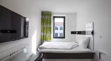 Ett kompakt modernt hotellrum med en enkelsäng, vita sängkläder och en platt-TV på väggen. Ett litet fönster med gröna och vita gardiner släpper in naturligt ljus. Ett skrivbord med glasskiva finns bredvid sängen.