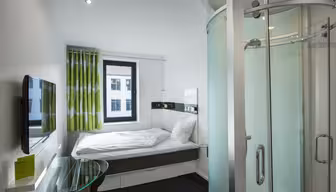 Ett kompakt och modernt hotellrum med en stor säng, en väggmonterad platt-TV, ett skrivbord i glas med en stol och en elegant duschvägg i böjd glas. Gröna och vita gardiner hänger vid fönstret och naturligt ljus fyller rummet.