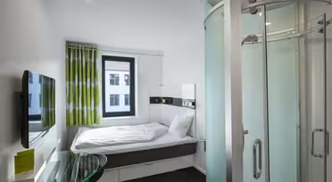 Ett kompakt och modernt hotellrum med en stor säng, en väggmonterad platt-TV, ett skrivbord i glas med en stol och en elegant duschvägg i böjd glas. Gröna och vita gardiner hänger vid fönstret och naturligt ljus fyller rummet.