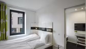 Ett modernt, minimalistiskt hotellrum med en snyggt bäddad säng, vita sängkläder och gardiner med gröna accenter. Ett stort fönster erbjuder utsikt över en grannbyggnad. En tv och ett skrivbord med en stol är synliga i ett angränsande rum.