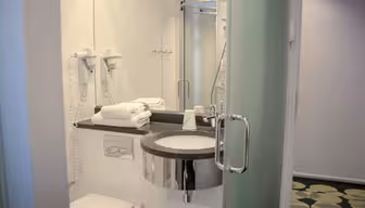 Modernt badrum med en väggmonterad hårtork, handfat med rund spegel, staplade handdukar och en inglasad dusch. Golvet har ett grönt cirkulärt mönster.