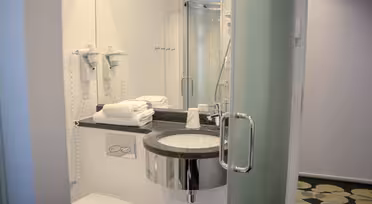 Modernt badrum med en väggmonterad hårtork, handfat med rund spegel, staplade handdukar och en inglasad dusch. Golvet har ett grönt cirkulärt mönster.