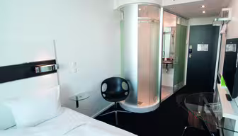 Modernt hotellrum med 1 enkelsäng, cirkulär glasdusch, svart stol, skrivbord i glas och platt-TV. Rummet har en modern design med skarpa linjer och en neutral färgpalett. Entrén och badrummet syns i bakgrunden.