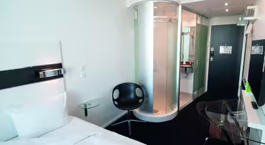 Modernt hotellrum med 1 enkelsäng, cirkulär glasdusch, svart stol, skrivbord i glas och platt-TV. Rummet har en modern design med skarpa linjer och en neutral färgpalett. Entrén och badrummet syns i bakgrunden.