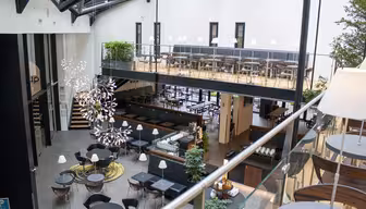 Rymligt, modernt inomhuscafé med entresolplan. På nedervåningen finns runda bord och stolar, och på övervåningen finns det fler sittplatser. Glasräcken och stora fönster ger naturligt ljus. Frodiga gröna växter och abstrakta ljuskronor pryder utrymmet.