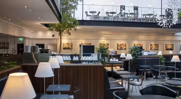 Modern caféinteriör med snygga svarta möbler, vita hängande lampor och gröna växter. En mezzanin med bord och stolar syns ovanför. Stora inramade foton kantar väggarna och ger rummet en modern touch.
