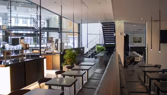 En modern caféinredning med eleganta svarta bord och stolar ordnade i rader. Stora fönster släpper in naturligt ljus. Trappor går upp i bakgrunden. Gröna växter ger en touch av färg till den minimalistiska designen.