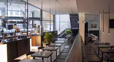 En modern caféinredning med eleganta svarta bord och stolar ordnade i rader. Stora fönster släpper in naturligt ljus. Trappor går upp i bakgrunden. Gröna växter ger en touch av färg till den minimalistiska designen.