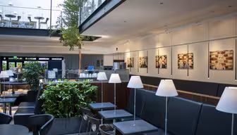 Modern caféinteriör med minimalistisk design, med svarta bord och stolar, vita hängande lampor och gröna växter. Stora fönster släpper in naturligt ljus. Fotografier pryder väggarna och en övre balkongyta är synlig.