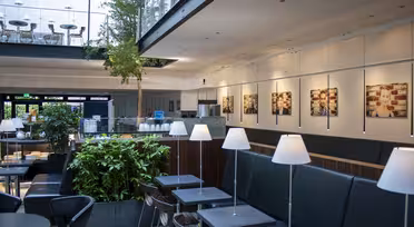 Modern caféinteriör med minimalistisk design, med svarta bord och stolar, vita hängande lampor och gröna växter. Stora fönster släpper in naturligt ljus. Fotografier pryder väggarna och en övre balkongyta är synlig.