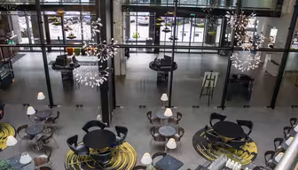 Ett modernt och luftigt café med runda bord och stolar uppställda på mönstrade mattor. Eleganta hängande armaturer ger en dekorativ touch. Stora glasfönster ger utsikt över parkerade bilar och gatan utanför.