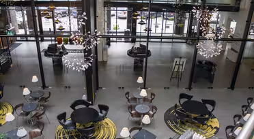 Ett modernt och luftigt café med runda bord och stolar uppställda på mönstrade mattor. Eleganta hängande armaturer ger en dekorativ touch. Stora glasfönster ger utsikt över parkerade bilar och gatan utanför.