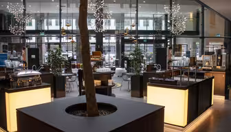 Modern caféinteriör med stora fönster och sittgrupper. En central plantering med ett träd är omgiven av upplysta serveringsstationer. Dekorativa armaturer hänger i taket och växter placeras runt utrymmet för stämning.