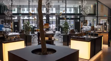 Modern caféinteriör med stora fönster och sittgrupper. En central plantering med ett träd är omgiven av upplysta serveringsstationer. Dekorativa armaturer hänger i taket och växter placeras runt utrymmet för stämning.