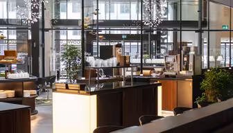 Modern caféinteriör med trädiskar och vita tallrikar. En kaffemaskin är synlig, omgiven av pallar. Stora glasfönster släpper in naturligt ljus, och dekorativa taklampor förstärker den eleganta atmosfären. Gröna växter ger en touch av naturen.