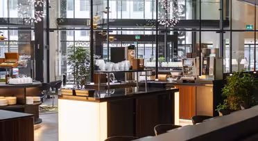 Modern caféinteriör med trädiskar och vita tallrikar. En kaffemaskin är synlig, omgiven av pallar. Stora glasfönster släpper in naturligt ljus, och dekorativa taklampor förstärker den eleganta atmosfären. Gröna växter ger en touch av naturen.