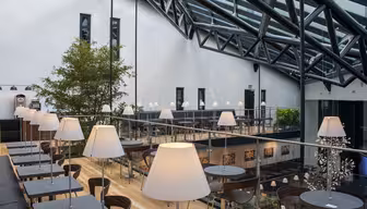 Modernt atrium med stort glastak, med ett träd, flera bord med lampor och trägolv. Utrymmet är uppdelat i två plan med sittgrupper och dekorativ konst på väggarna.