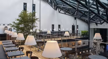 Modernt atrium med stort glastak, med ett träd, flera bord med lampor och trägolv. Utrymmet är uppdelat i två plan med sittgrupper och dekorativ konst på väggarna.