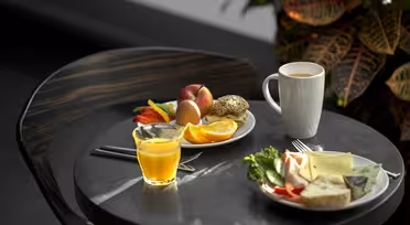 Frukost på ett runt bord med en mugg kaffe, ett glas apelsinjuice, en tallrik med olika frukter och en tallrik med bröd, ost och grönsaker. En krukväxt syns i bakgrunden.