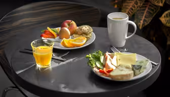 Ett runt bord med ett frukostpåslag inklusive apelsinjuice, en kaffe i en vit mugg, en tallrik med frukt, bröd, ett ägg och en annan tallrik med ost, sallad och en croissant. En växt syns i bakgrunden.