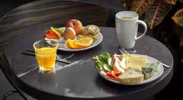 Ett runt bord med ett frukostpåslag inklusive apelsinjuice, en kaffe i en vit mugg, en tallrik med frukt, bröd, ett ägg och en annan tallrik med ost, sallad och en croissant. En växt syns i bakgrunden.