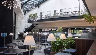 Modern caféinteriör med högt i tak och takfönster. Sittdelen på övervåningen har bord och stolar, medan nedervåningen har svarta bord, vita lampor och krukväxter. Unika hängande armaturer lägger till dekoration.