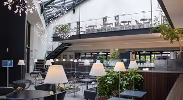 Modern caféinteriör med högt i tak och takfönster. Sittdelen på övervåningen har bord och stolar, medan nedervåningen har svarta bord, vita lampor och krukväxter. Unika hängande armaturer lägger till dekoration.