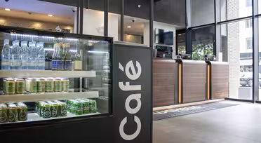Modern kaféinteriör med en glasmonter full av vatten på flaska och burkar till vänster. En svart caféskylt syns och disken med träpaneler i bakgrunden. Stora fönster släpper in naturligt ljus utifrån.