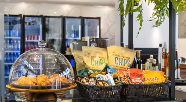 En cafédisk med en mängd olika snacks. Det finns croissanter under en glaskupol, korgar fyllda med chips, jordnötter och Haribo-gummi. Dryckskylar finns i bakgrunden och gröna växter hänger ovanför.