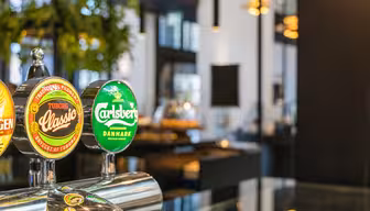 Närbild på ölkranar med etiketter för Tuborg Classic och Carlsberg på en bar. Bakgrunden är mjukt suddig och visar inomhusväxter och modern inredning med varm belysning.