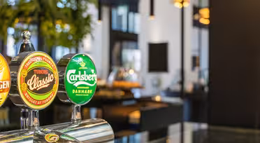 Närbild på ölkranar med etiketter för Tuborg Classic och Carlsberg på en bar. Bakgrunden är mjukt suddig och visar inomhusväxter och modern inredning med varm belysning.