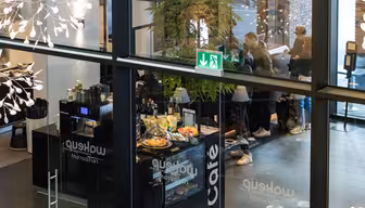 Se genom glas av ett livligt kafé med folk i kö, en disk med bakverk och drycker och dekorativa lampor som hänger. En utgångsskylt syns ovanför entrén och en stor växt ger utrymmet grönska.