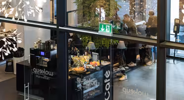 Se genom glas av ett livligt kafé med folk i kö, en disk med bakverk och drycker och dekorativa lampor som hänger. En utgångsskylt syns ovanför entrén och en stor växt ger utrymmet grönska.