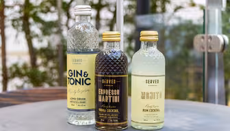 Tre flaskor SERVADE cocktails står uppställda på ett bord. Smakerna inkluderar Gin & Tonic, Espresso Martini och Mojito. De har färgglada etiketter och är placerade mot en suddig utomhusbakgrund med grönska.