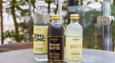 Tre flaskor SERVADE cocktails står uppställda på ett bord. Smakerna inkluderar Gin & Tonic, Espresso Martini och Mojito. De har färgglada etiketter och är placerade mot en suddig utomhusbakgrund med grönska.