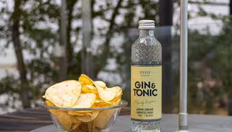 En glasskål med potatischips står på ett bord bredvid en drink på flaska märkt "Gin & Tonic". Bakgrunden presenterar grönt lövverk och en metallstolpe. Scenen föreslår en utomhusmiljö.